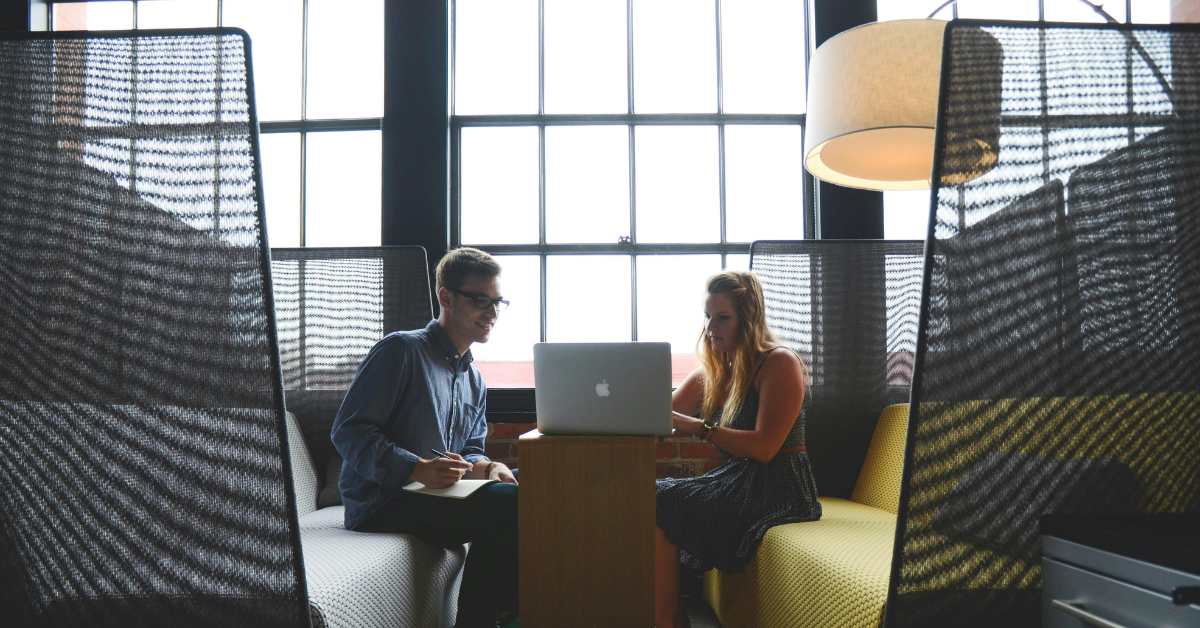 Founder and team member collaborating in a booth, as the woman types on a laptop and the man smiles while taking notes, illustrating trust, teamwork, and vulnerability in leadership.